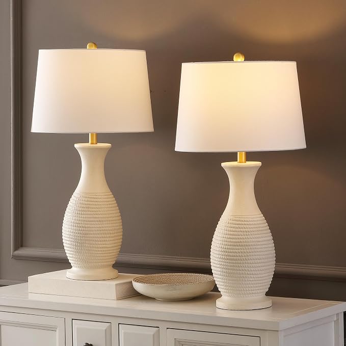 Two matching table lamps with textured cream ceramic bases and white drum shades, lit and displayed on a white cabinet against a brown wall.